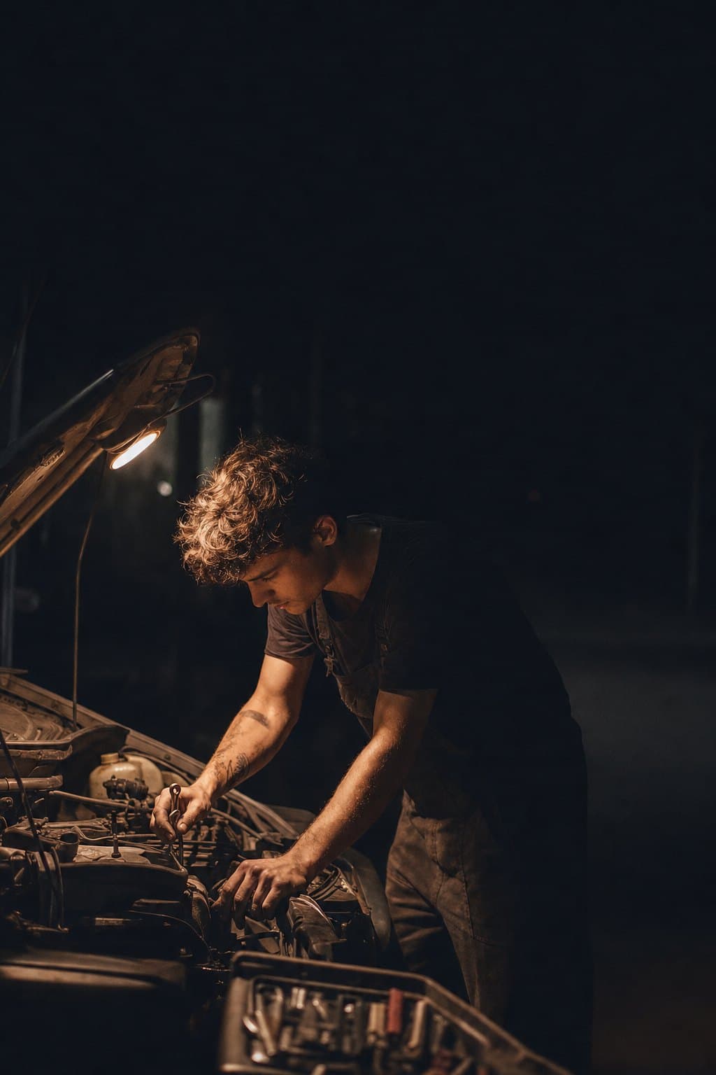 A mechanic leaning into an engine bay under a work light at night